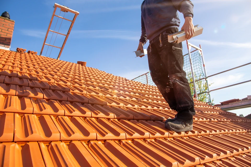Photographie en extérieur d'un chantier de rénovation de toiture. Un ouvrier en tenue professionnelle travaille sur une toiture en cours de réparation ou de remplacement, visible sur la structure d'un bâtiment. La scène montre clairement des travaux de couverture toiture (tuiles, ardoise ou matériau de couverture), avec un environnement de chantier organisé et professionnel en pleine lumière naturelle.